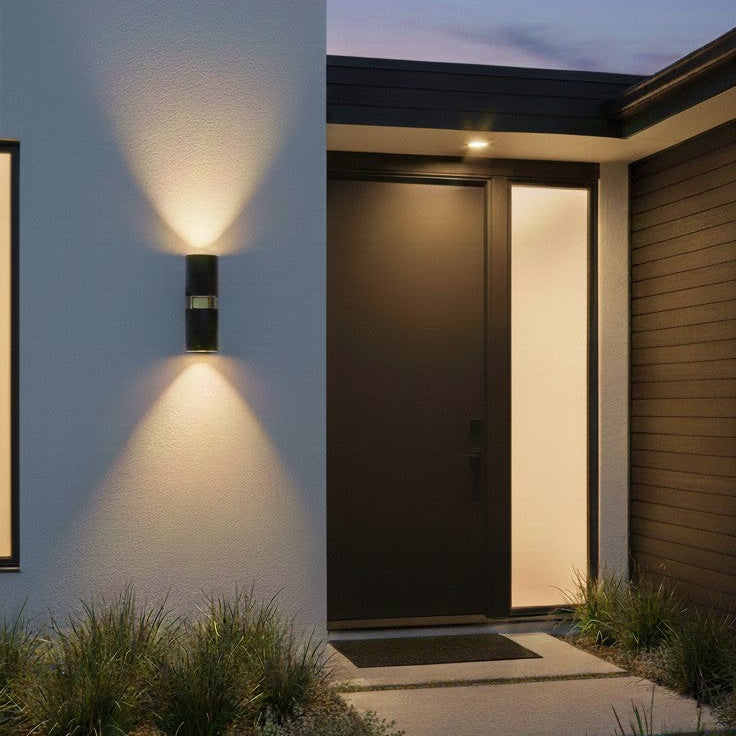 Black cylindrical outdoor wall light illuminating a modern home entrance at dusk