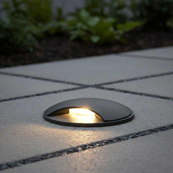 Round ground light with a dark grey finish, illuminating a paved path