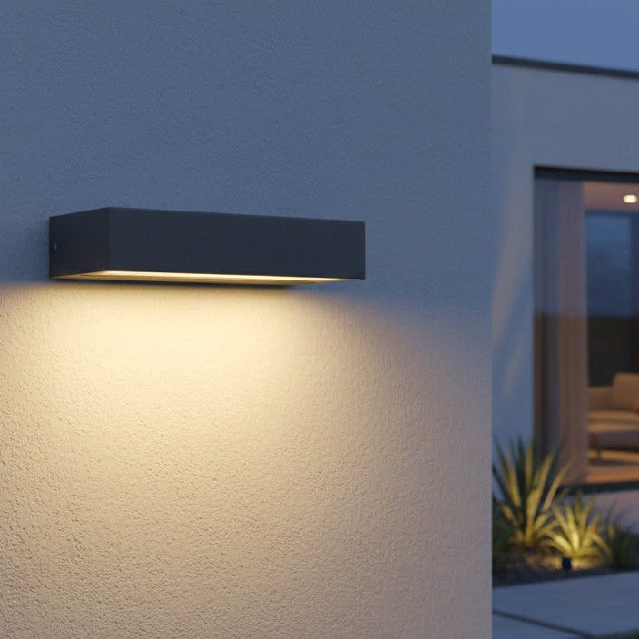 Rectangular outdoor wall light in dark grey illuminates a textured wall at dusk