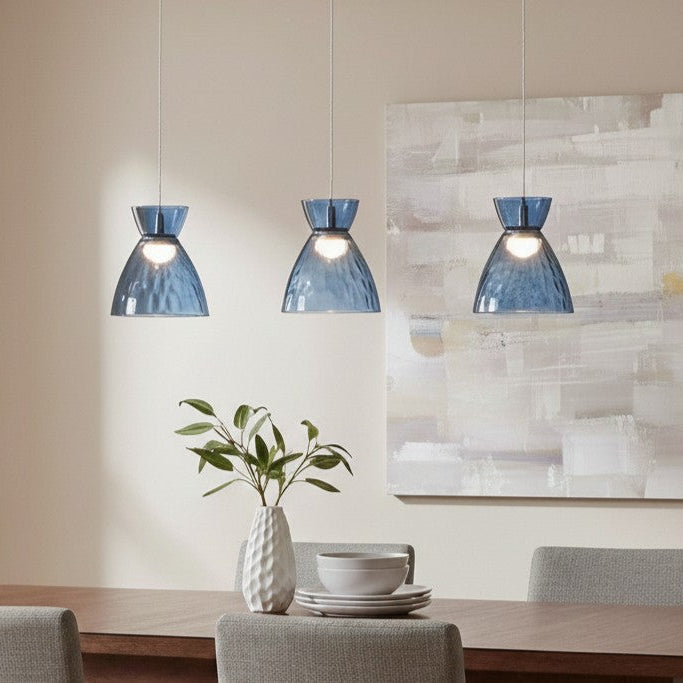 Three blue glass pendant lights hang above a dining table with a white vase and dishes