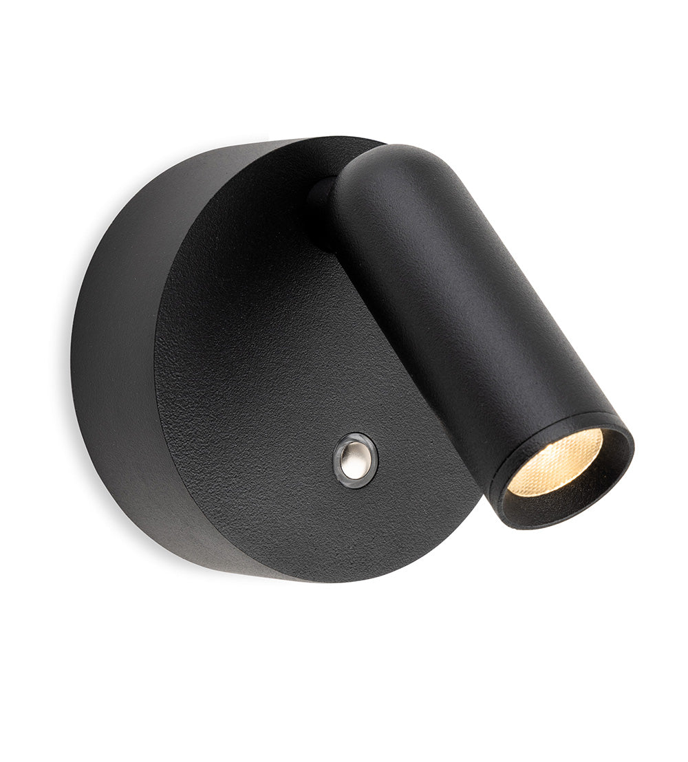 Black wall-mounted spotlight with a circular base and an adjustable cylindrical head, emitting a warm light