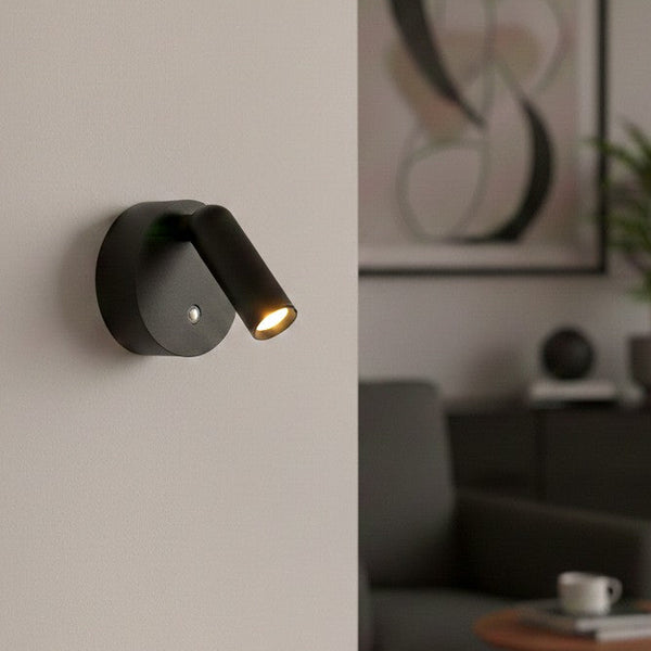 Black wall-mounted spotlight illuminates a light gray wall, with a blurred living room in the background