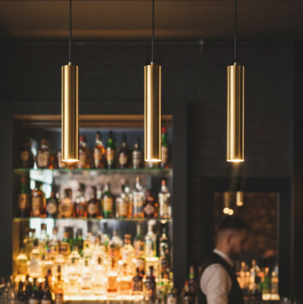 Three gold cylindrical pendant lights illuminate a bar with bottles on shelves and a bartender