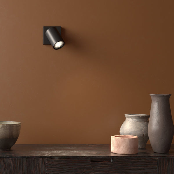 Black cylindrical wall light on a brown wall, illuminating decorative pottery on a dark wooden surface