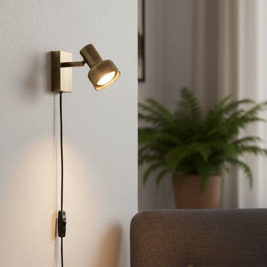 Brass wall light with a black cord and switch, illuminating a white wall next to a green plant