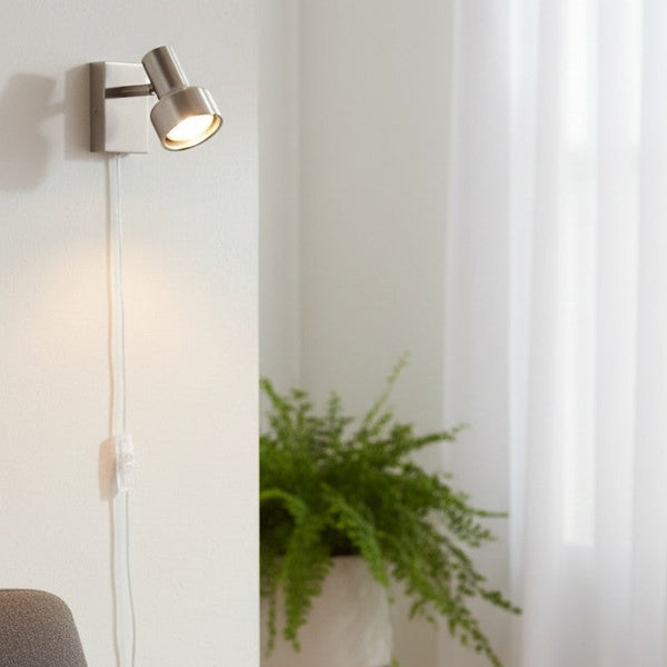 Brushed metal wall-mounted spotlight with a white cord and switch, illuminating a white wall above a green plant
