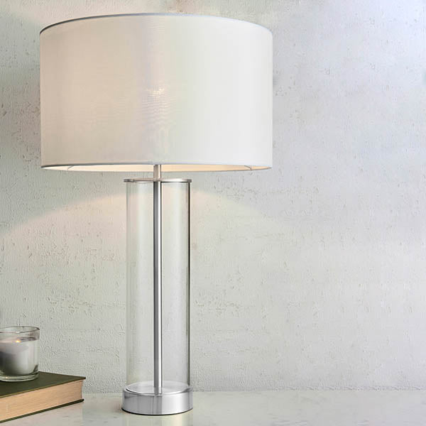 Table lamp with white shade, clear glass column, and brushed steel base, next to a book and candle.