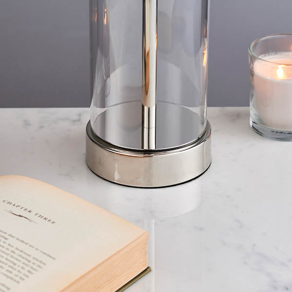 Close-up of the brushed steel base and clear glass column of a table lamp, with a book and candle.