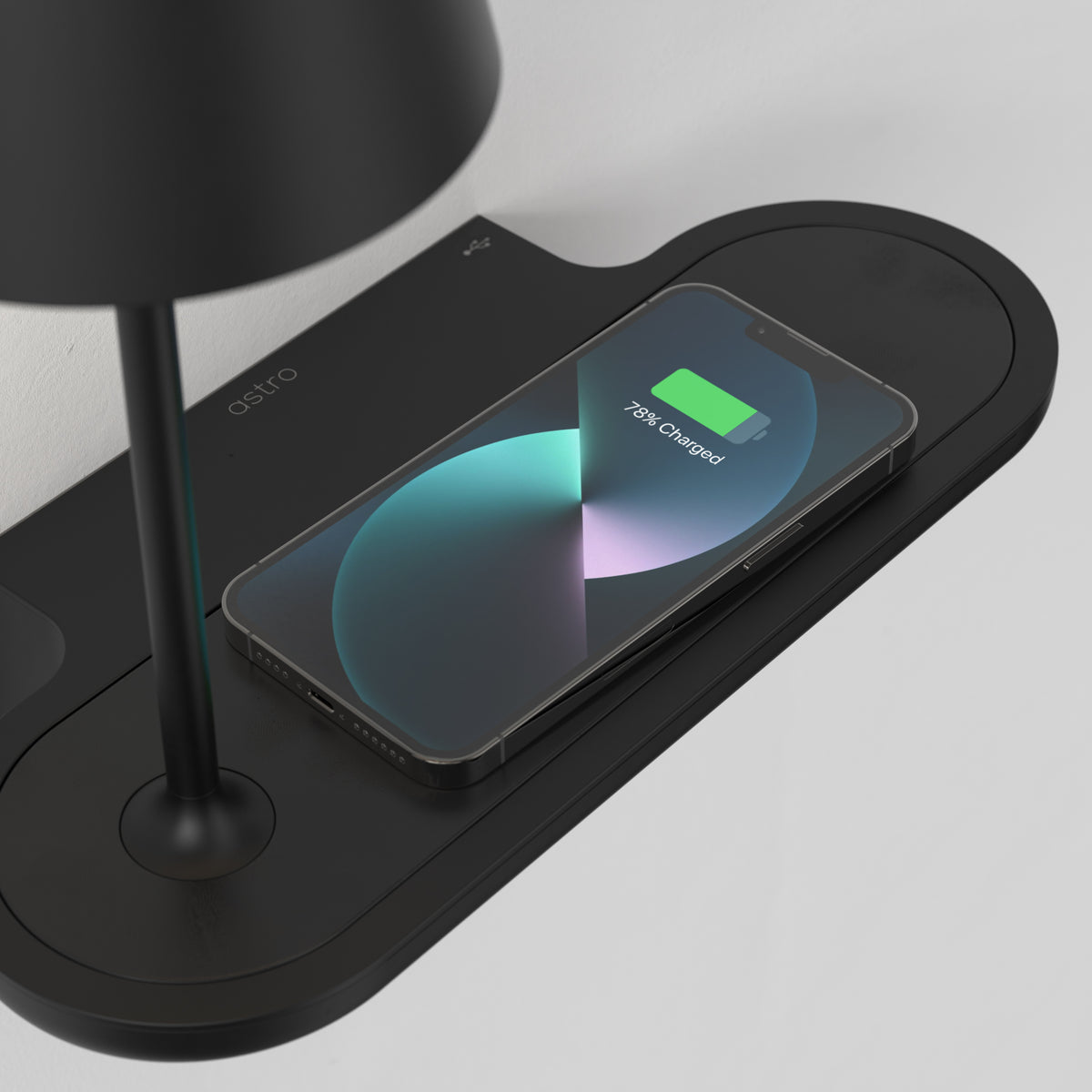 Black iPhone charging on a black wireless charging pad with a black lamp base next to it