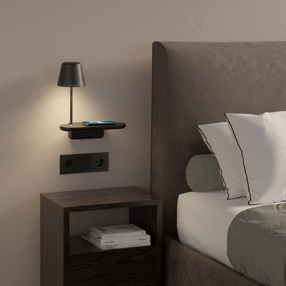 A bedside table with a lamp, phone, and books next to a bed with pillows