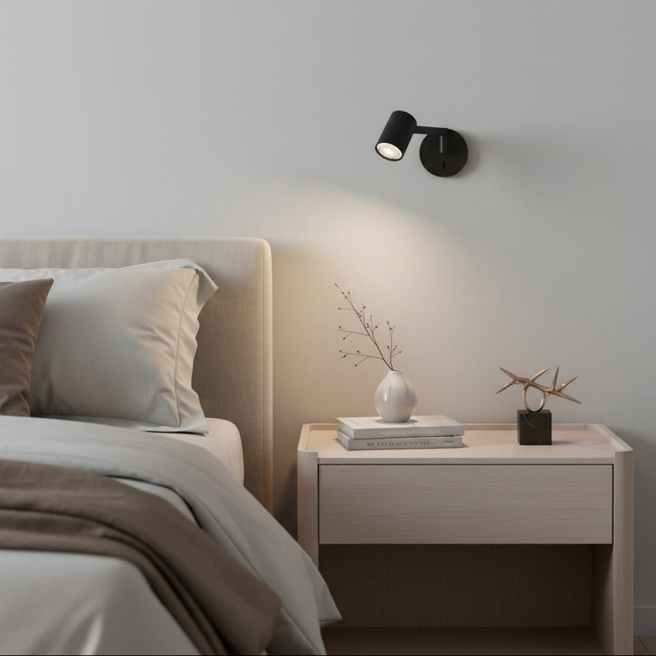 Black wall-mounted spotlight above a light wood bedside table with books and a vase