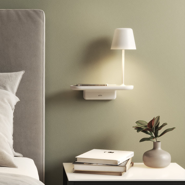 A white wall-mounted shelf with a lamp and phone, next to a bed with a grey headboard and a plant