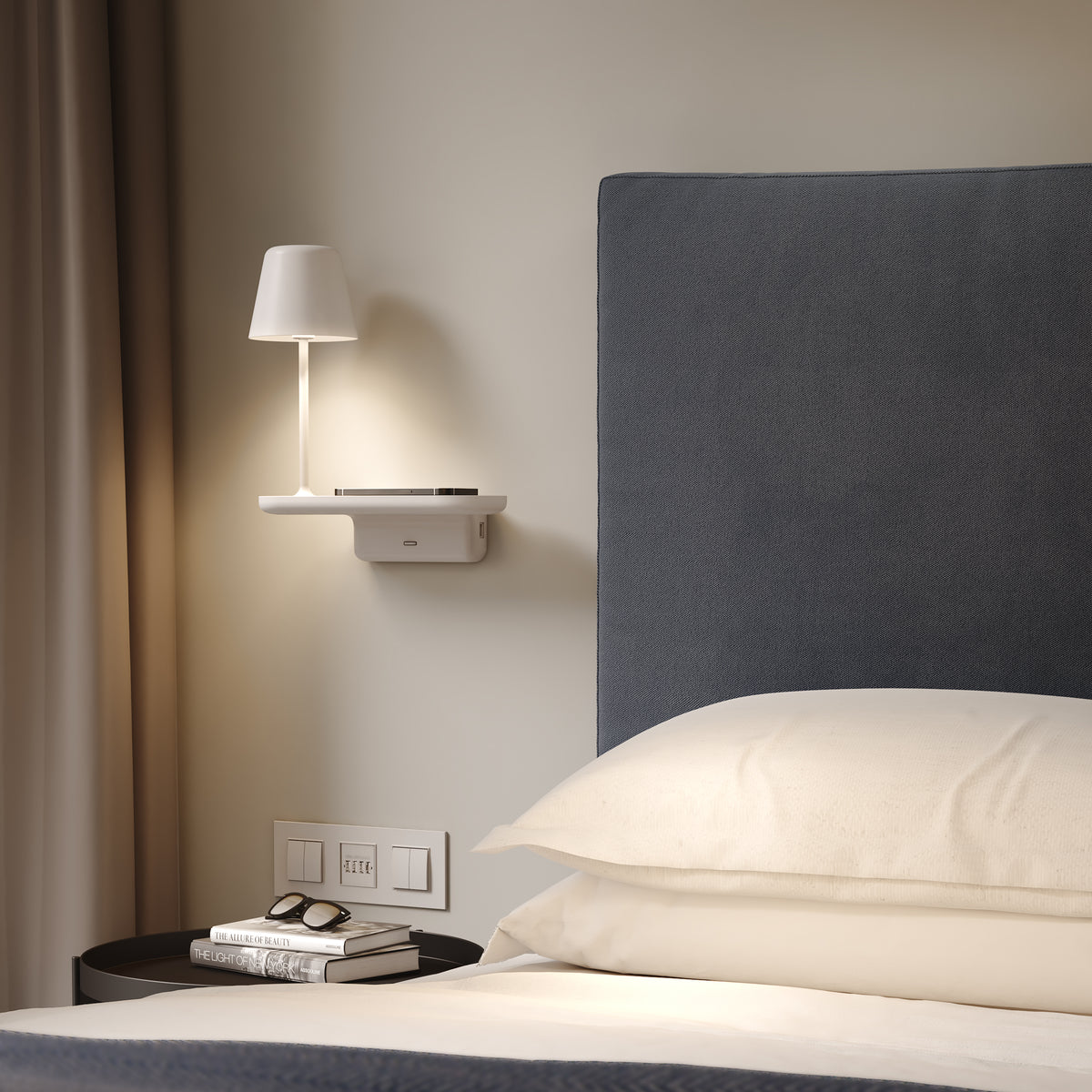 White bedside lamp with a phone charging on a shelf above a bed with white pillows and a blue headboard