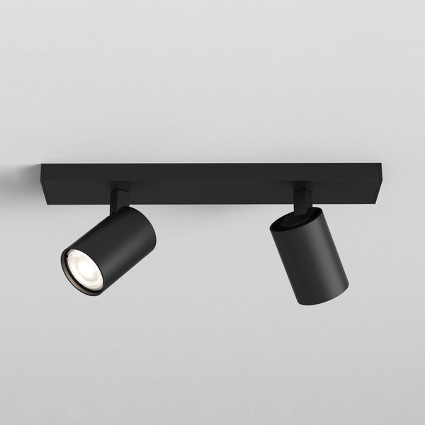Two black cylindrical spotlights on a black rectangular bar, one illuminated, against a white ceiling