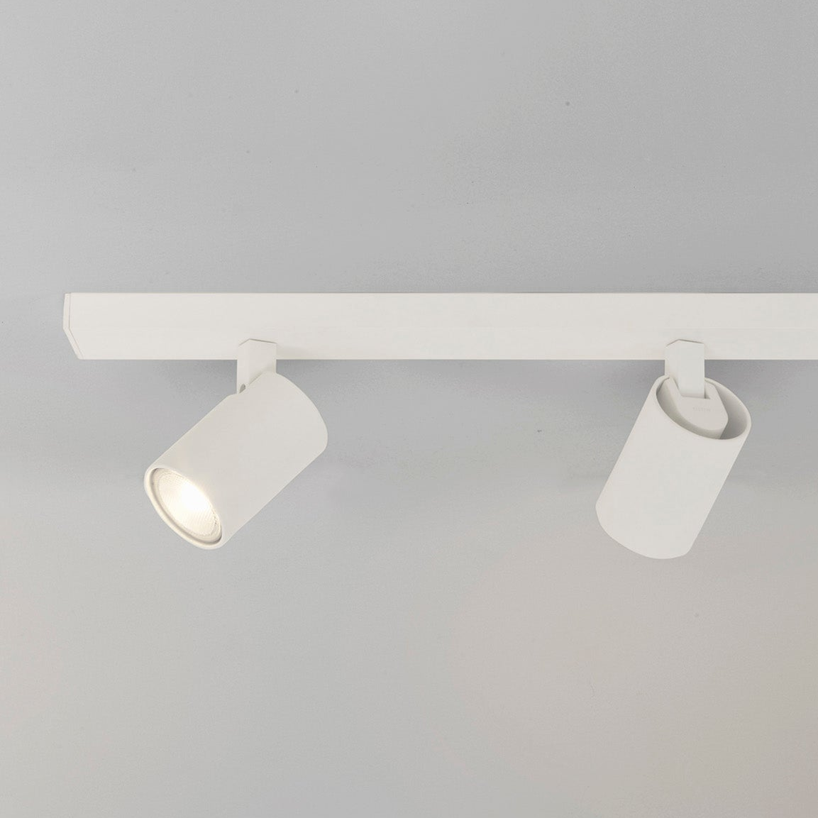 Two white cylindrical track lights on a white track, one illuminated