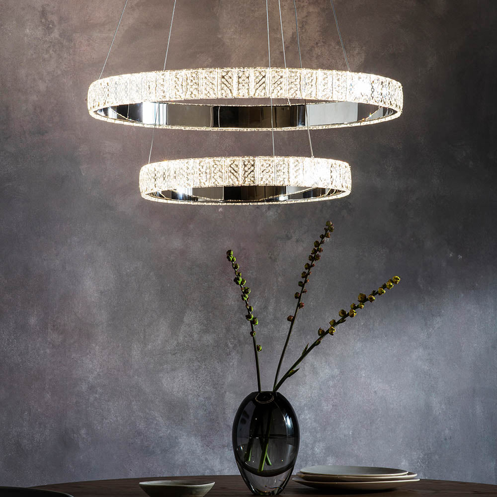 Two illuminated crystal ring chandeliers hang above a dark table with a vase and plates.