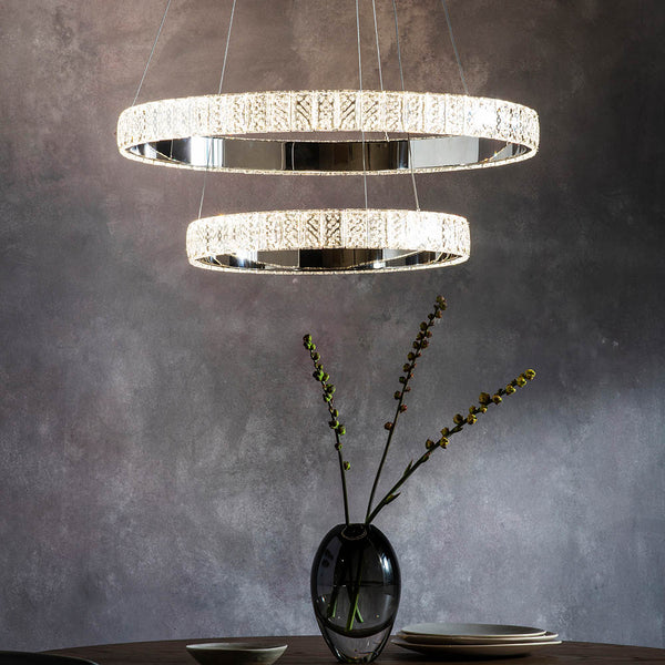 Two illuminated crystal ring chandeliers hang above a dark table with a vase and plates.