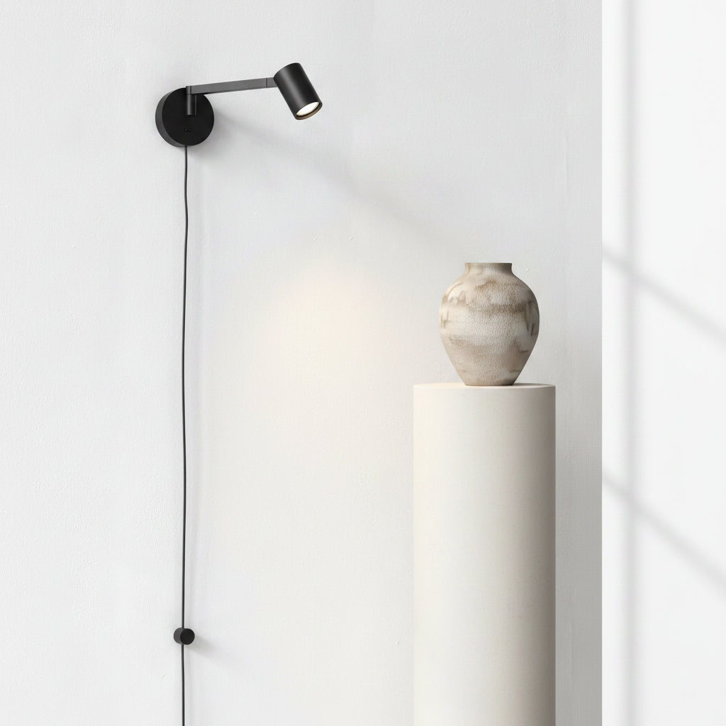 Black wall-mounted spotlight illuminating a textured vase on a white pedestal