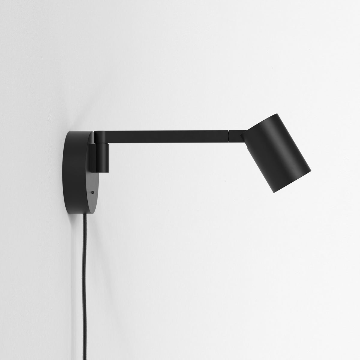 Black wall-mounted lamp with an adjustable arm and a cylindrical shade, against a white wall
