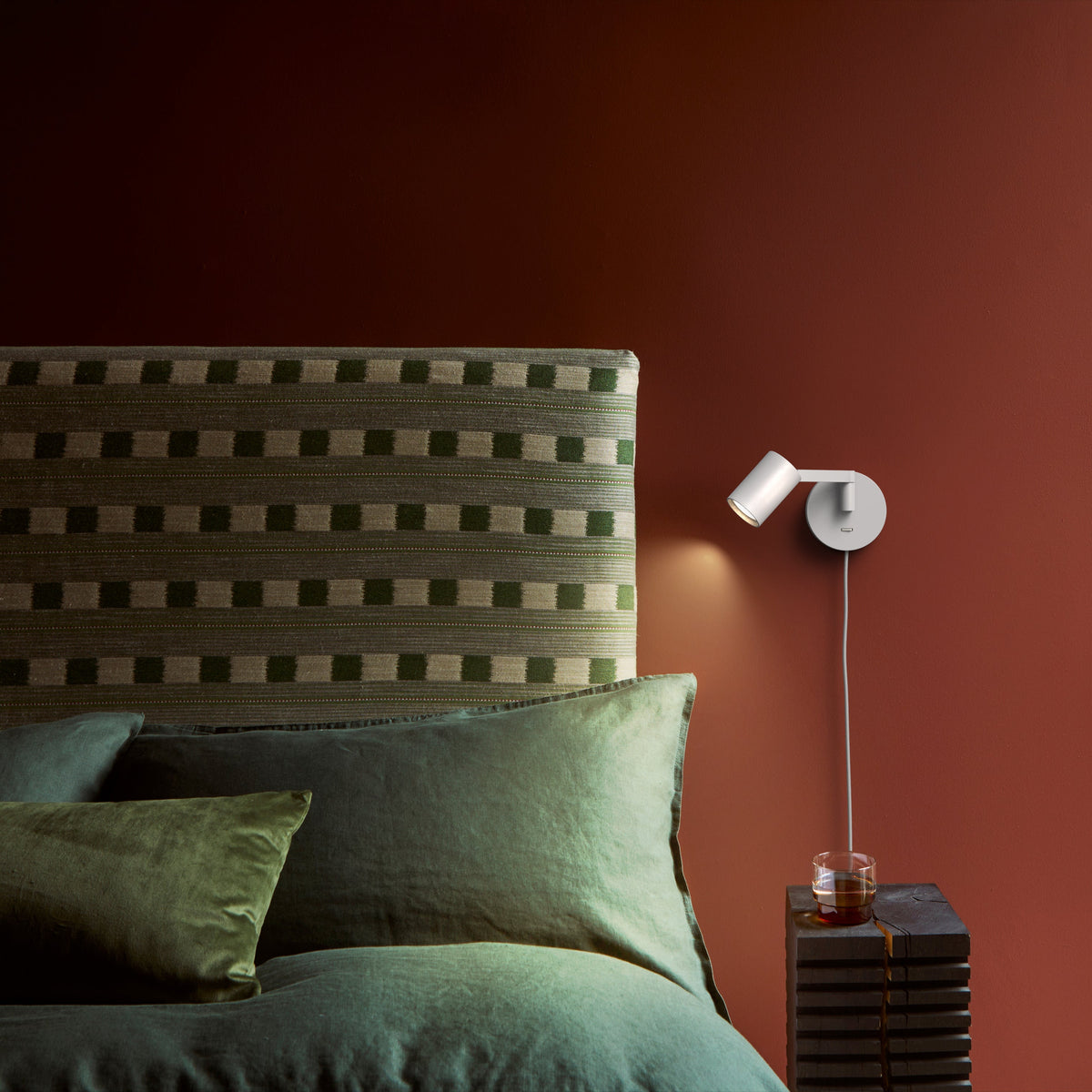 Bedroom with green patterned headboard, green pillows, white wall lamp, and a drink on a dark wooden side table