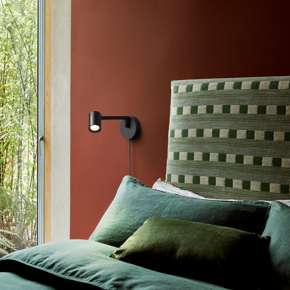 Black wall-mounted reading light above a bed with green pillows and a patterned headboard