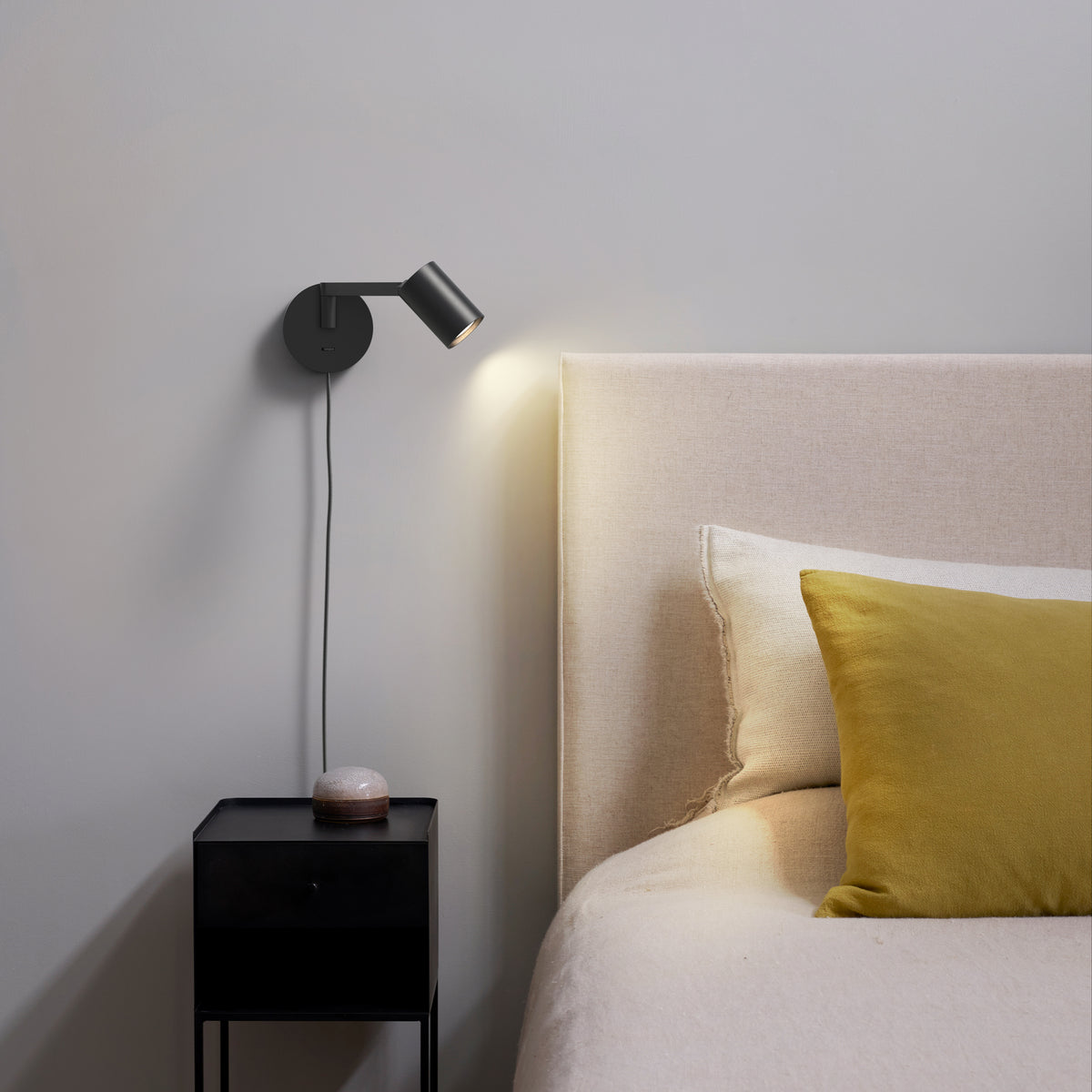 Black wall-mounted spotlight illuminates a bedroom with a beige headboard and a mustard yellow pillow