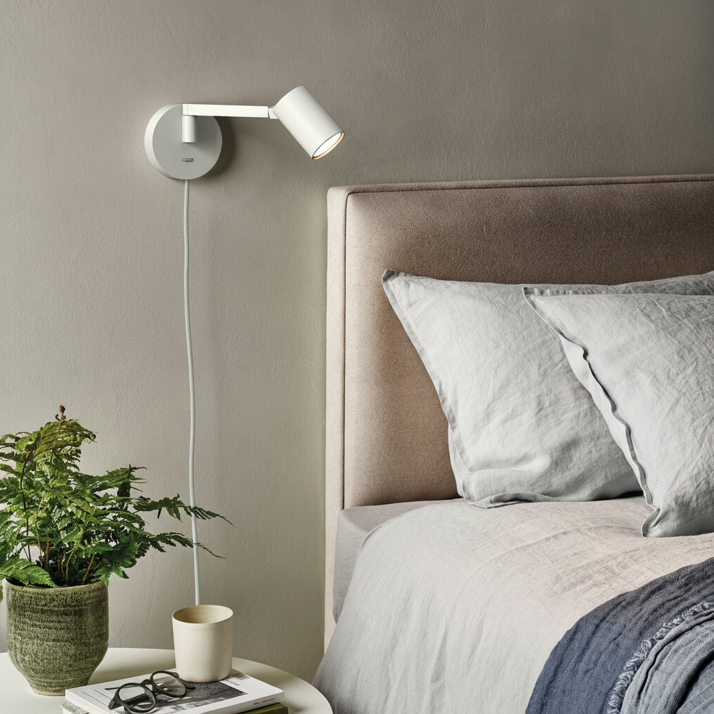 White wall-mounted reading light above a bed with light grey pillows and a blue throw blanket