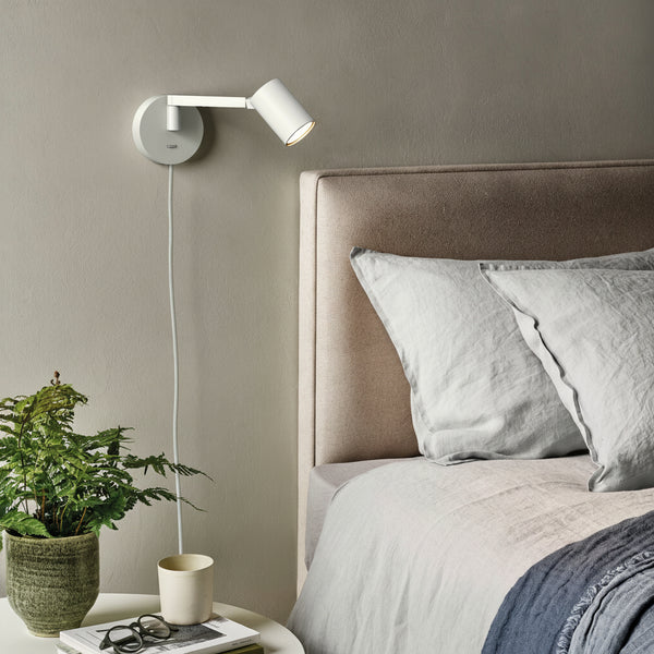 White wall-mounted reading light above a bed with light grey pillows and a blue throw blanket