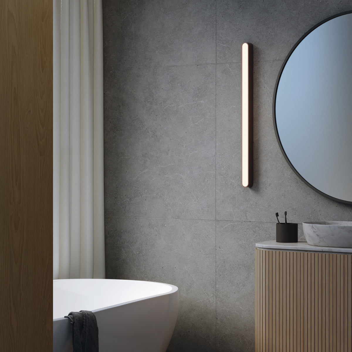 Bathroom with white tub, wood vanity, round mirror, and vertical light fixture on a gray tiled wall