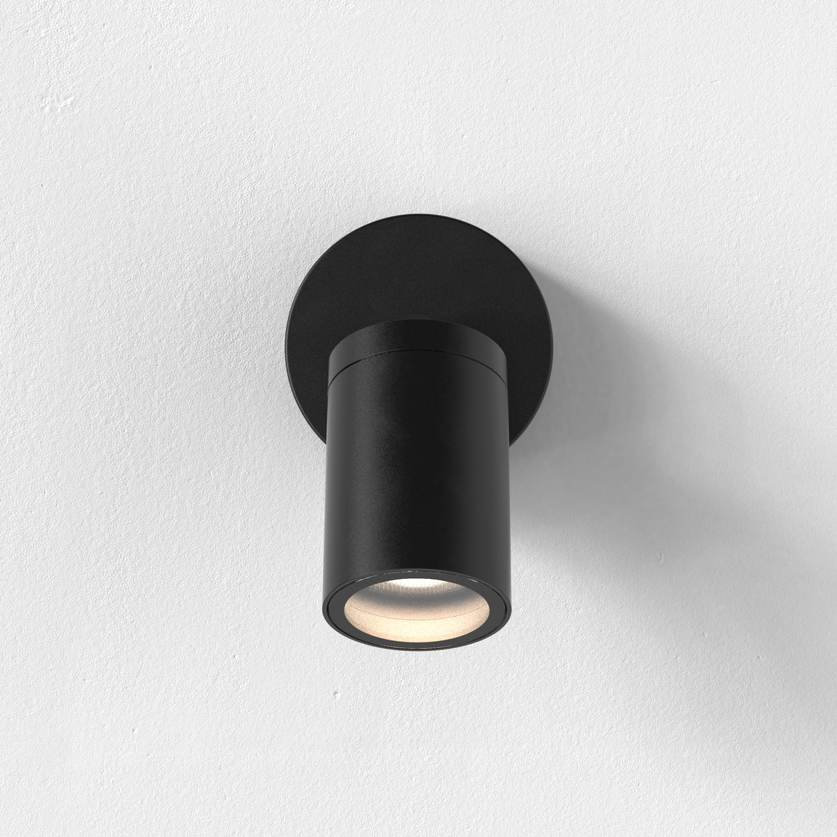 Black cylindrical wall light with a circular base, emitting a warm glow downwards onto a white textured wall