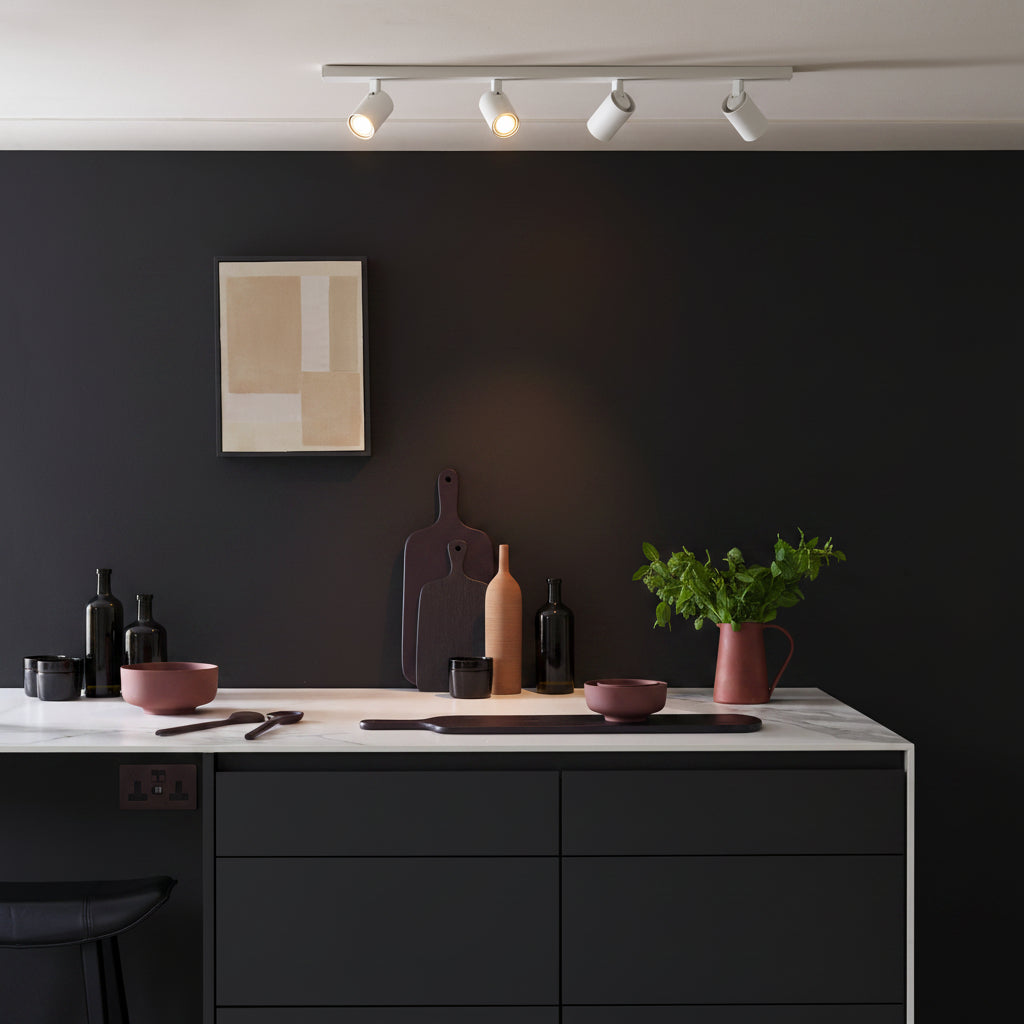 Four white track lights illuminate a dark kitchen with a marble countertop and black cabinets