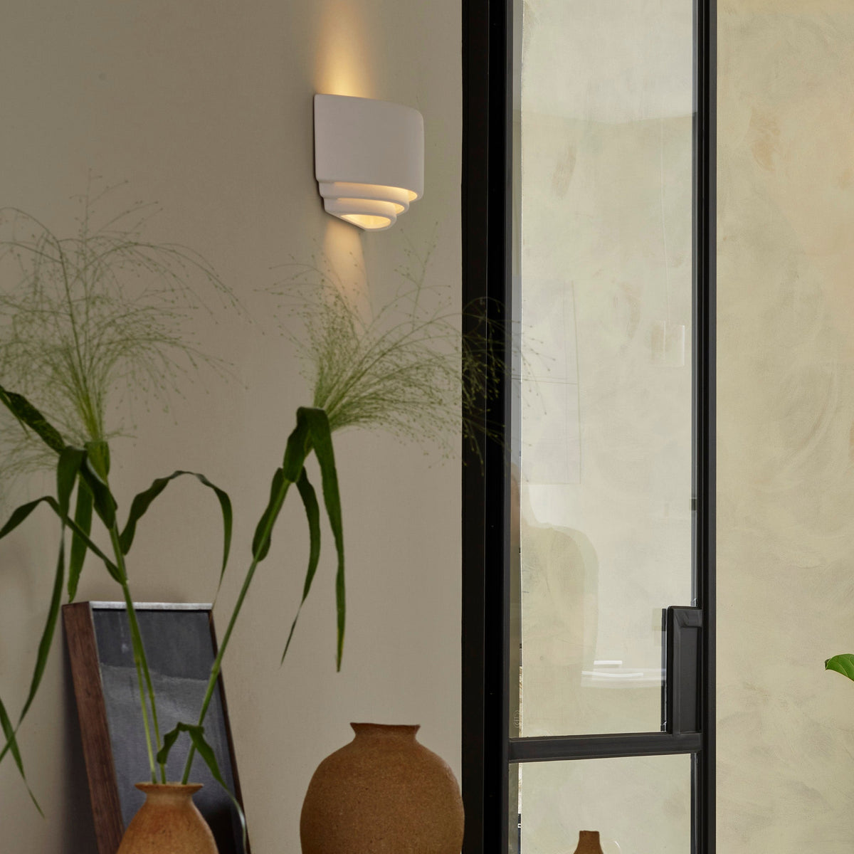 White wall sconce with tiered design casting light upwards and downwards, next to a black-framed glass door