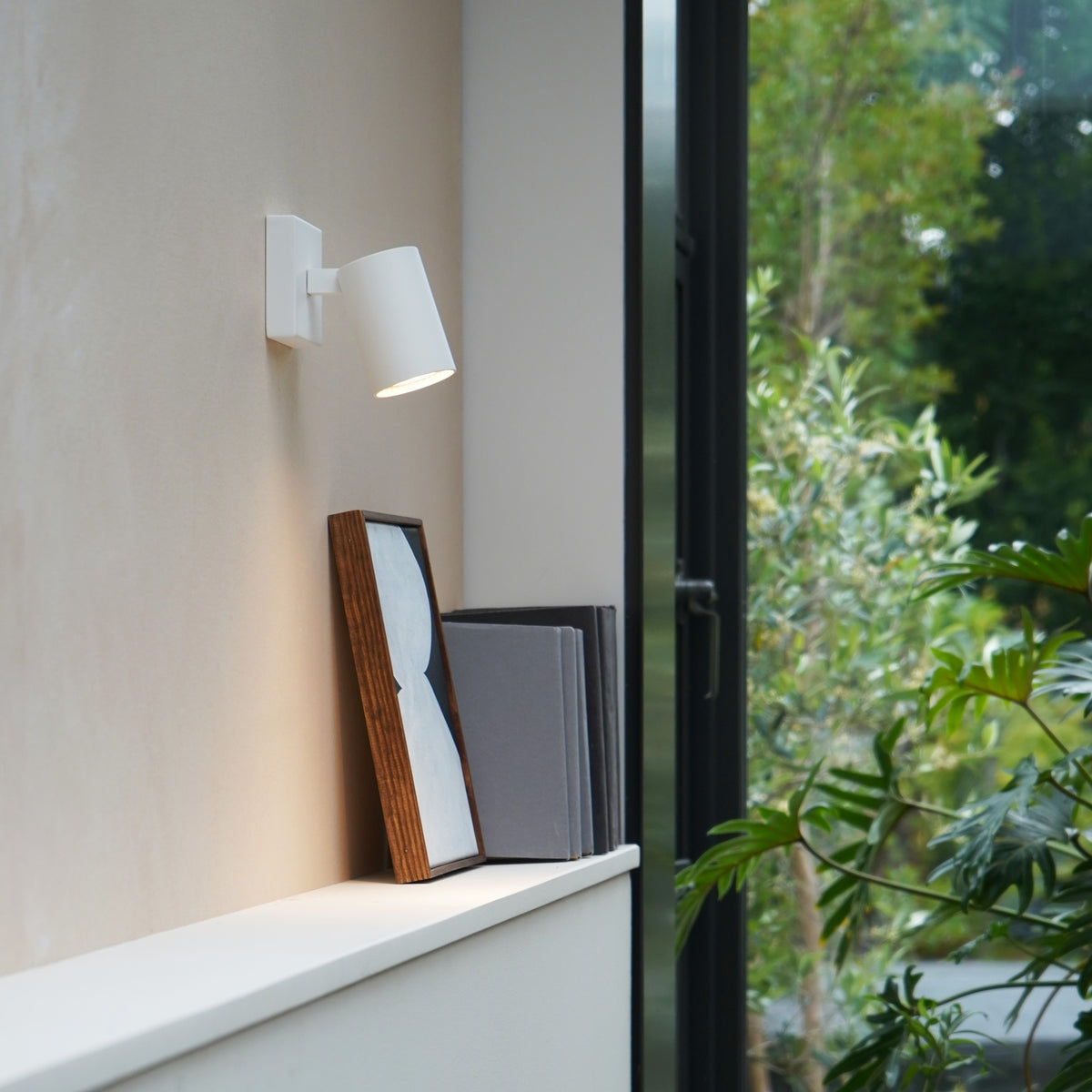 White wall light illuminating a framed picture and books on a shelf next to a window with green plants outside