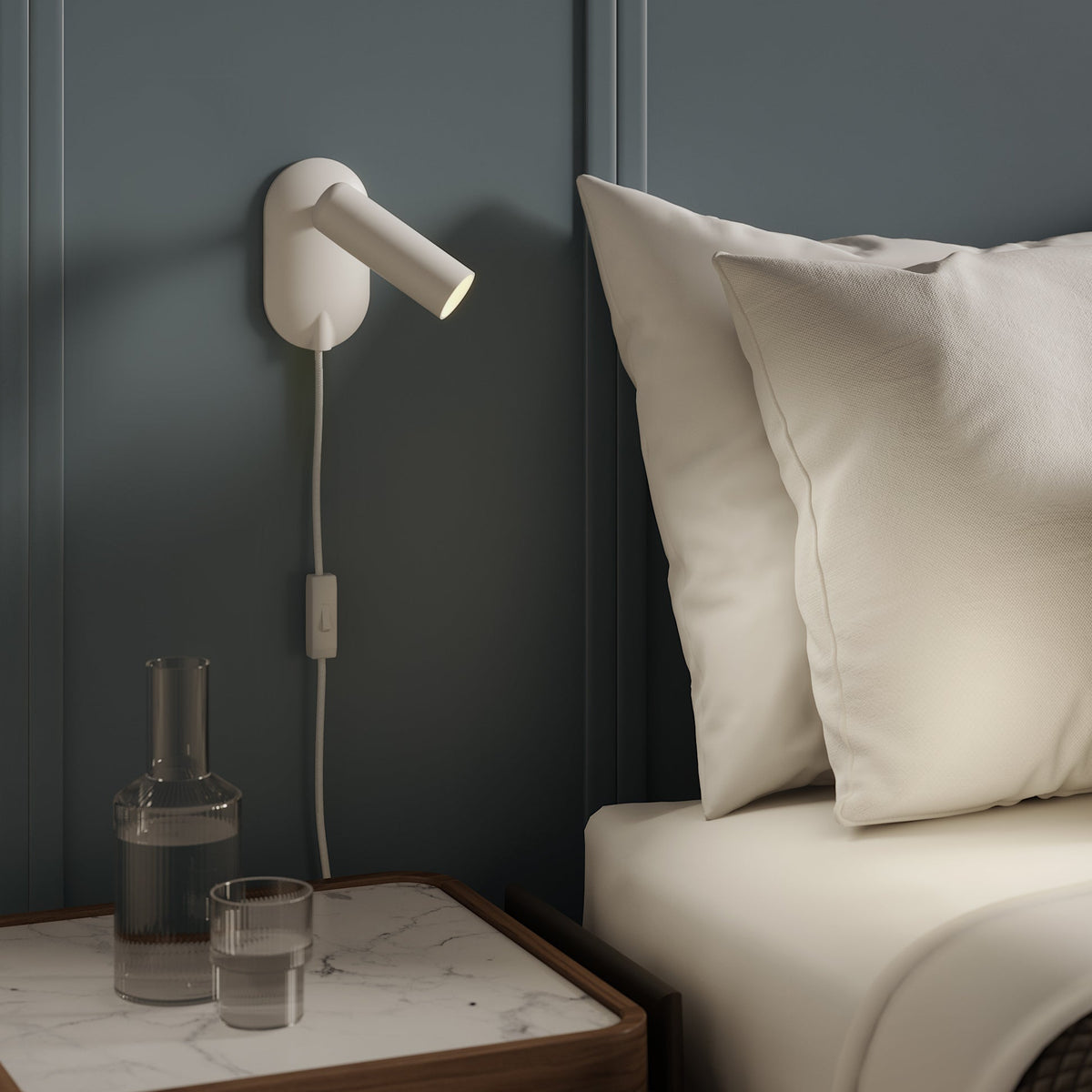 White wall-mounted reading light with a switch, next to a bed with white pillows and a marble-top nightstand