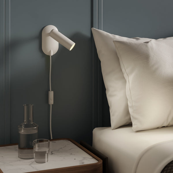 White wall-mounted reading light with a switch, next to a bed with white pillows and a marble-top nightstand