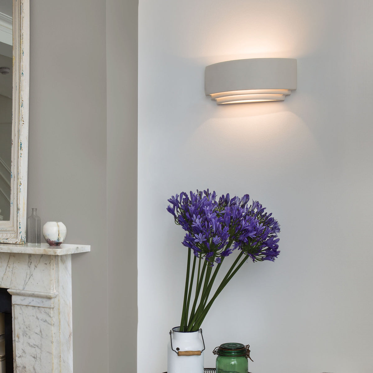 White wall light with curved layers, illuminating a wall above a vase of purple flowers