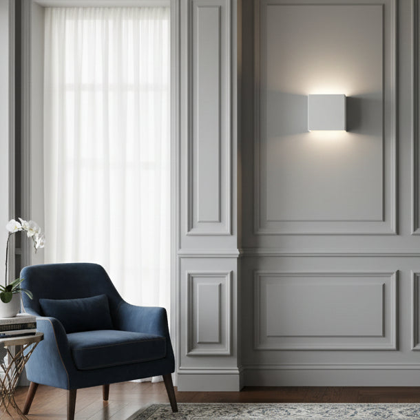 Square white wall light on a grey paneled wall above a blue armchair