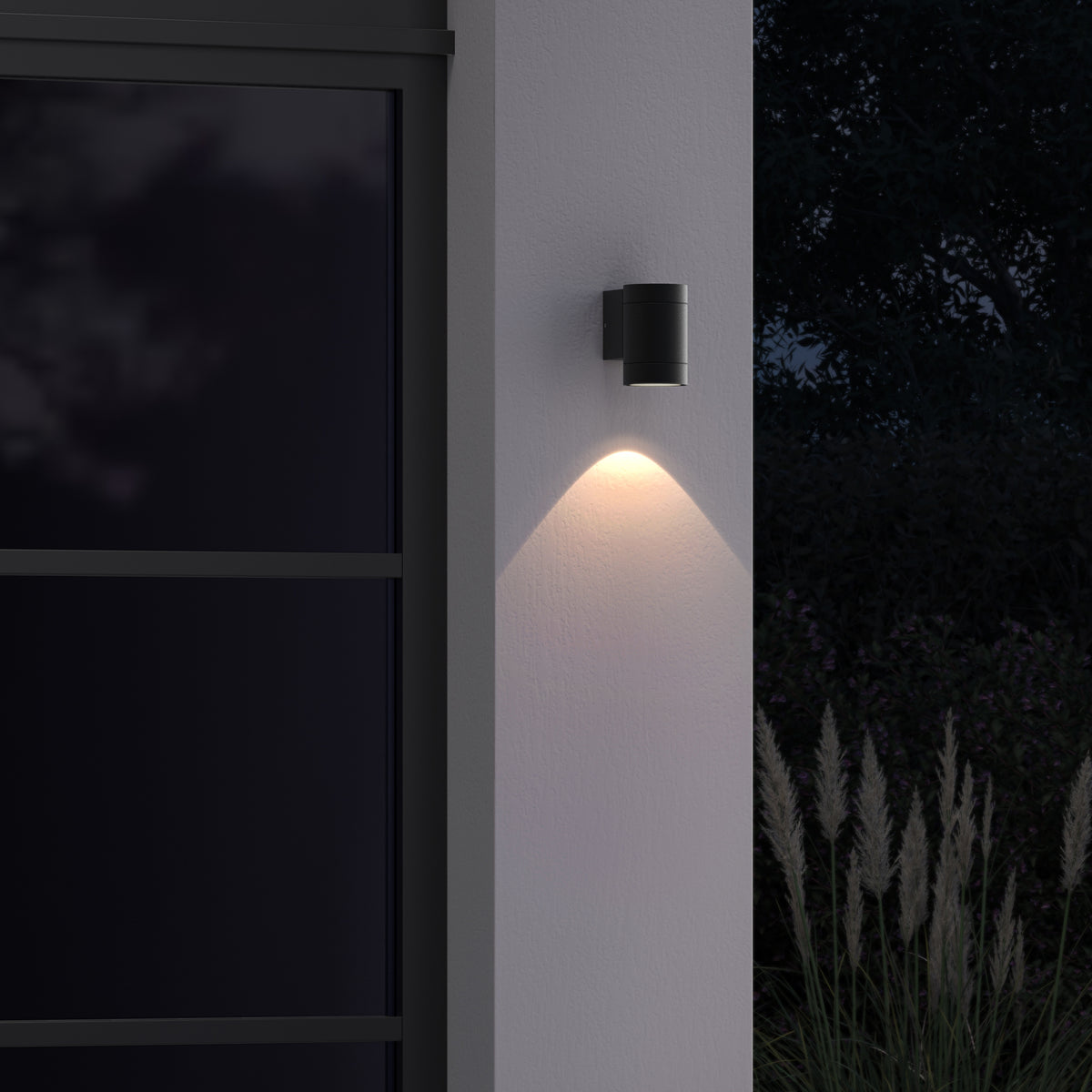 Black cylindrical outdoor wall light illuminating a white wall at night