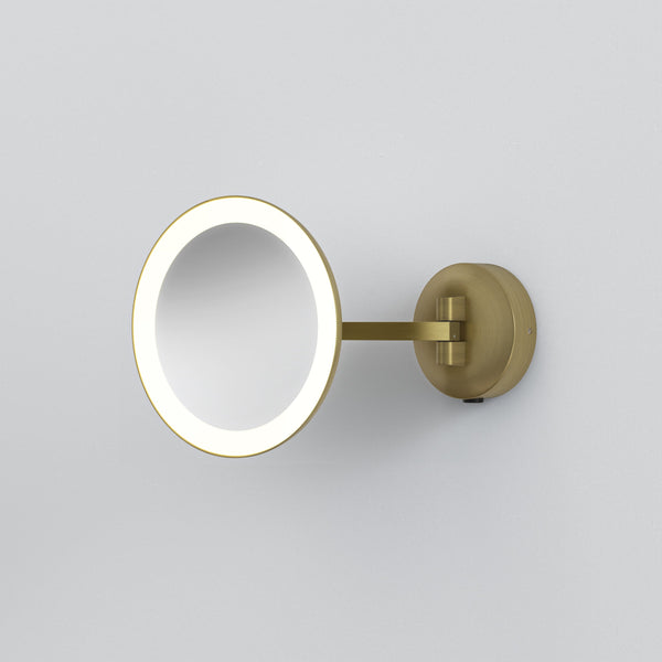 Round illuminated magnifying mirror with a brass finish, mounted on a white wall
