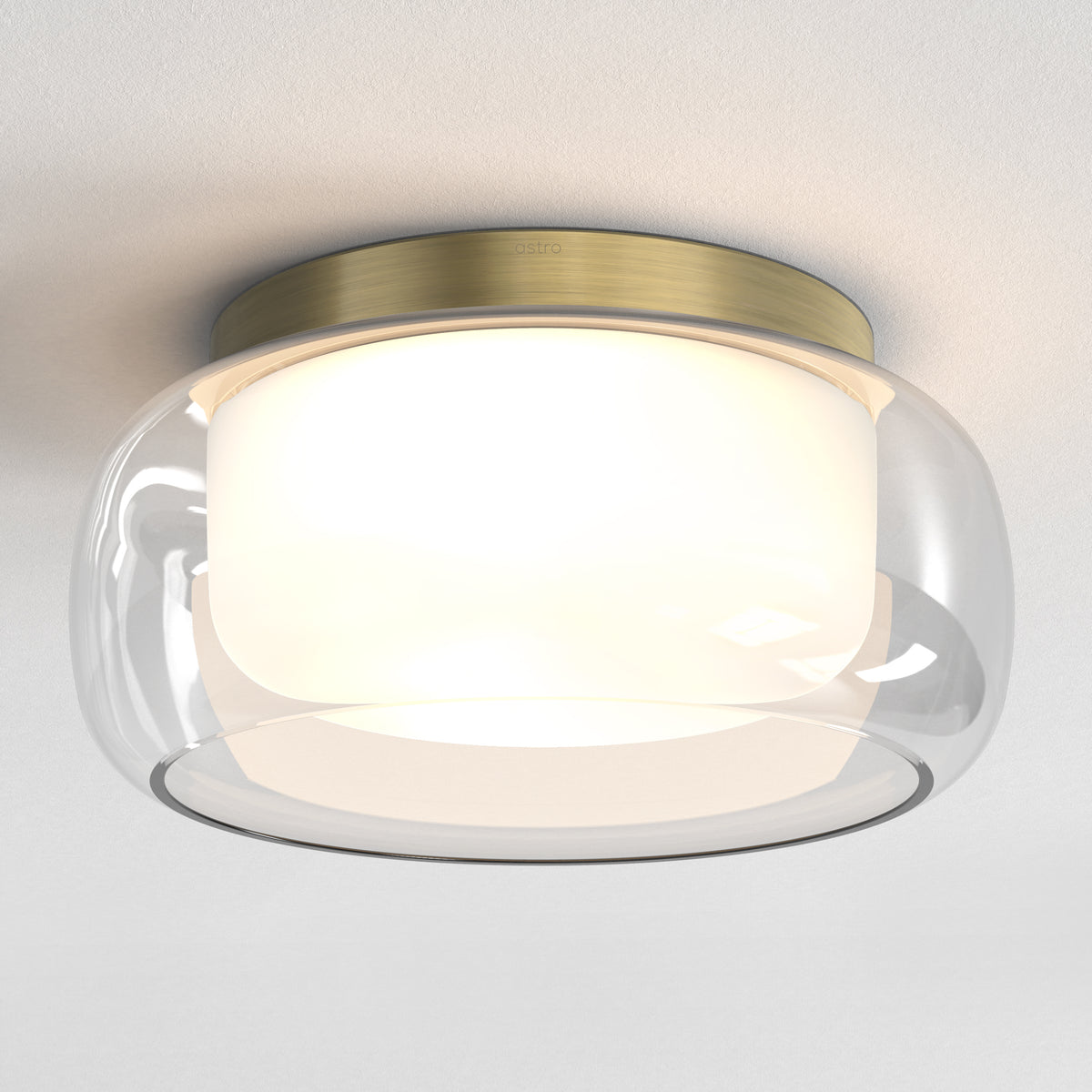 Astro ceiling light with a brass base, clear glass dome, and an inner white diffuser, illuminated on a white ceiling
