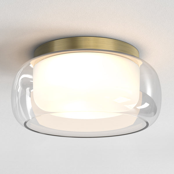 Astro ceiling light with a brass base, clear glass dome, and an inner white diffuser, illuminated on a white ceiling