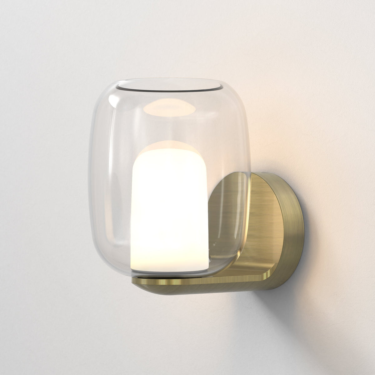 Clear glass wall sconce with a brushed brass base and a glowing white light source