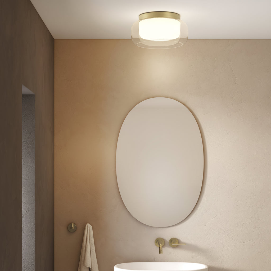 A modern bathroom with an oval mirror, a white sink, and a brass ceiling light