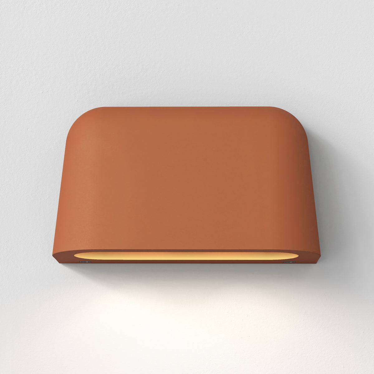 Terracotta wall light with rounded edges, casting a warm glow downwards onto a white wall
