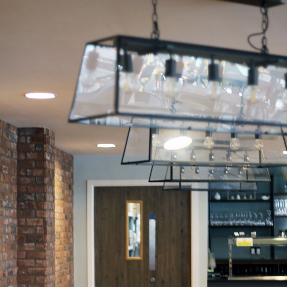 Industrial-style chandelier with exposed bulbs hangs over a modern bar area with brick walls and recessed lighting.