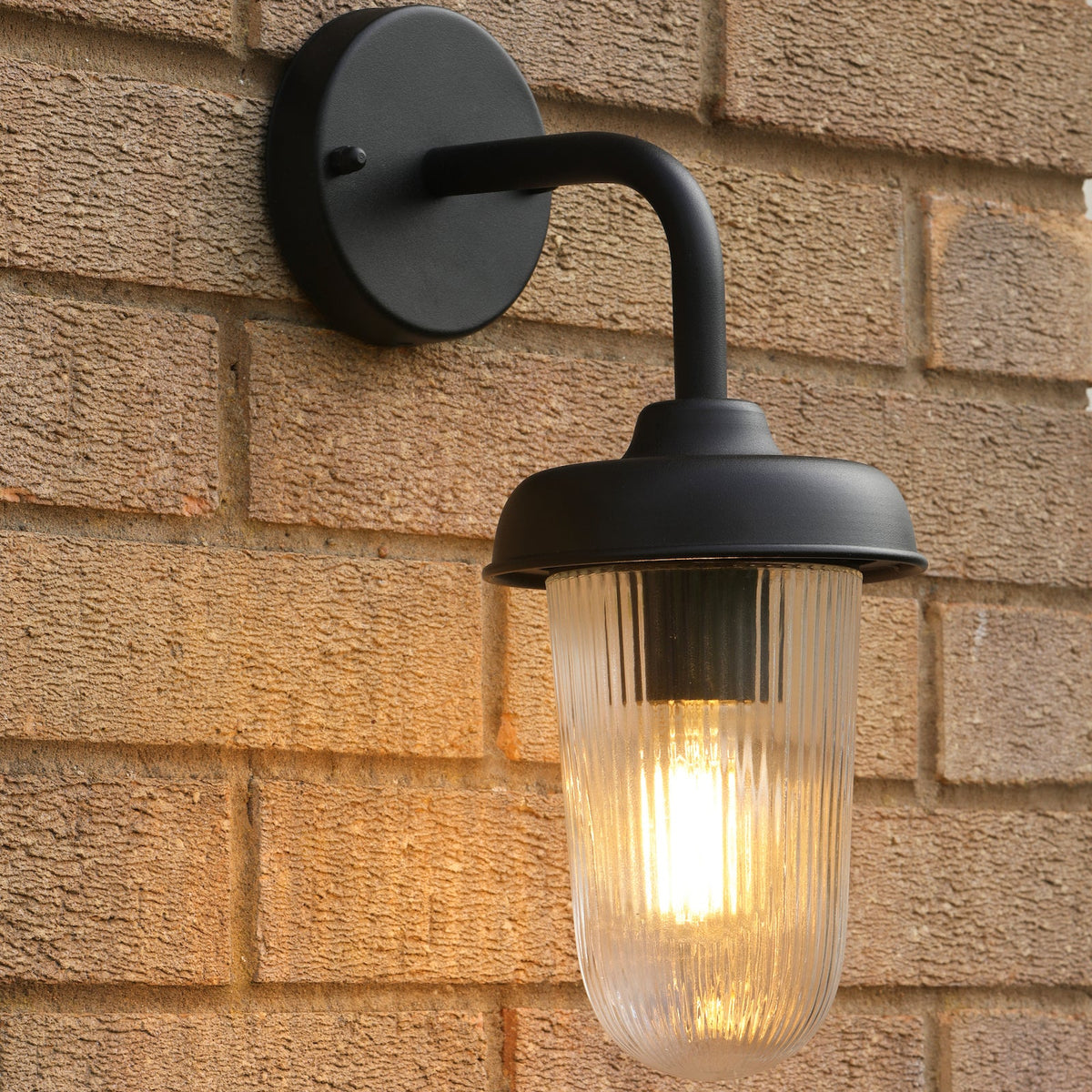 Black outdoor wall light with a ribbed glass shade and a warm glowing bulb against a brick wall