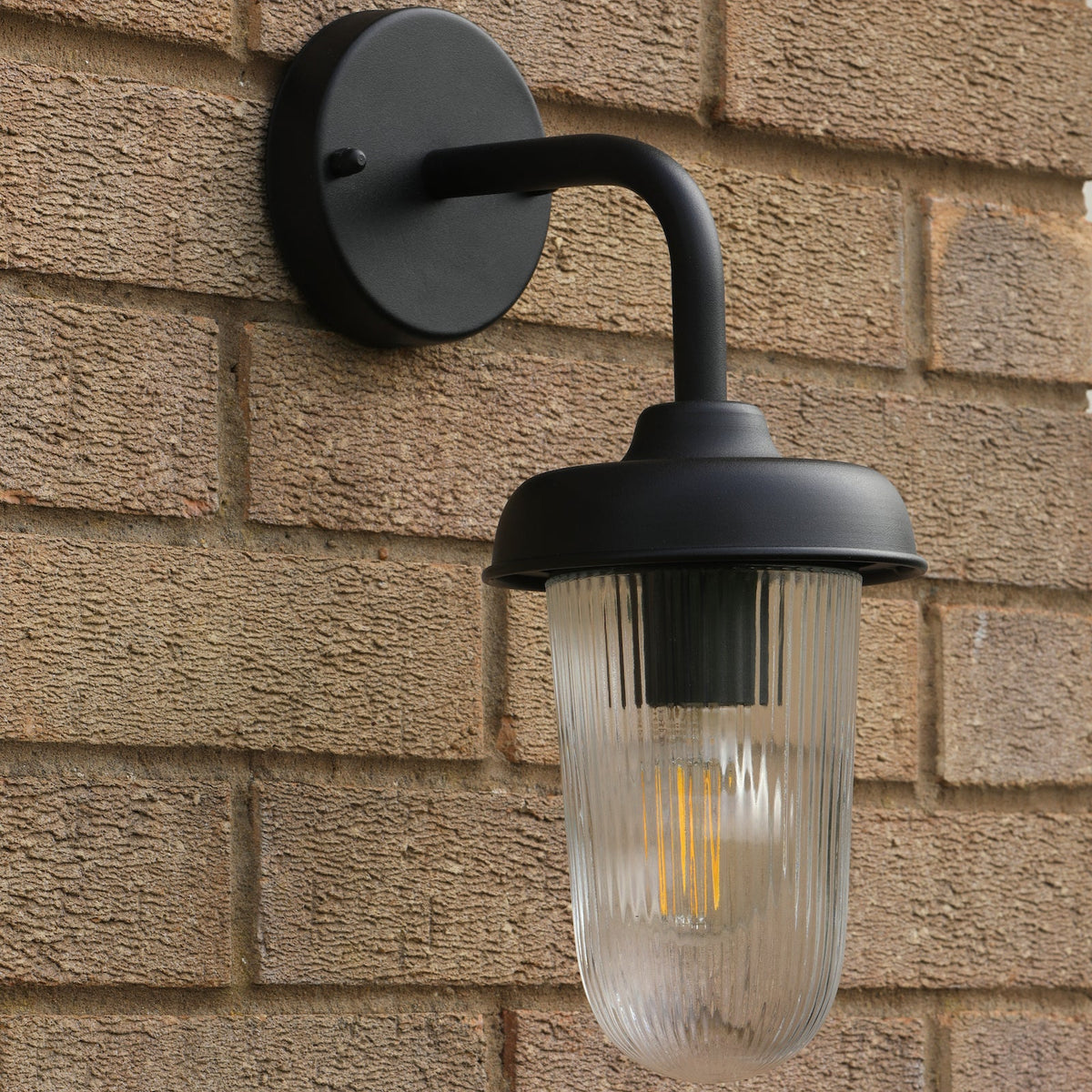Black outdoor wall light with a ribbed glass shade and a visible filament bulb on a brick wall