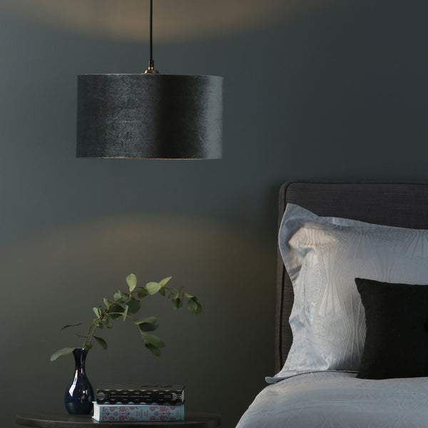 A dark gray velvet drum pendant light hangs above a nightstand with a blue vase, books, and a bed.