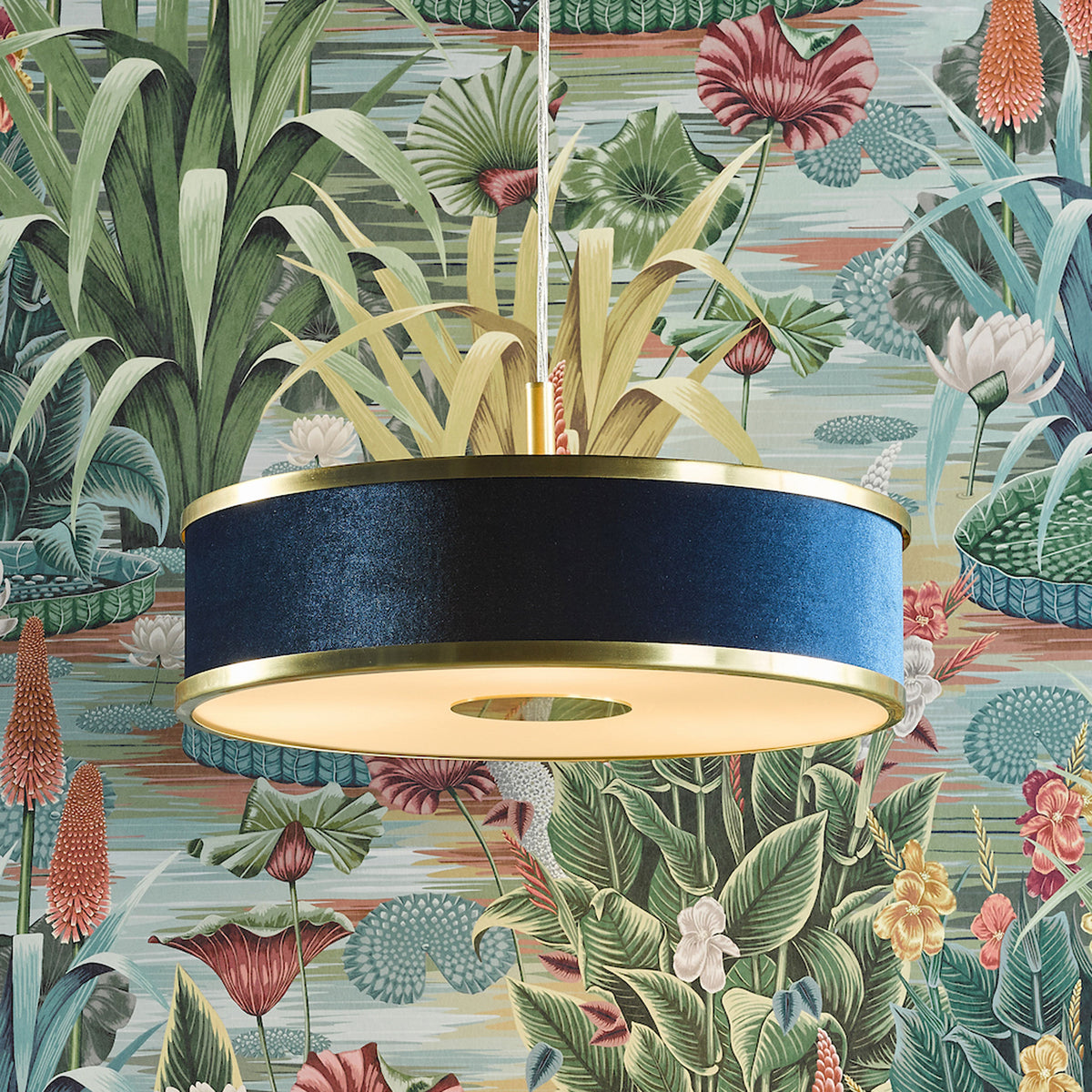 Blue velvet drum pendant light with brass accents against a vibrant botanical wallpaper.