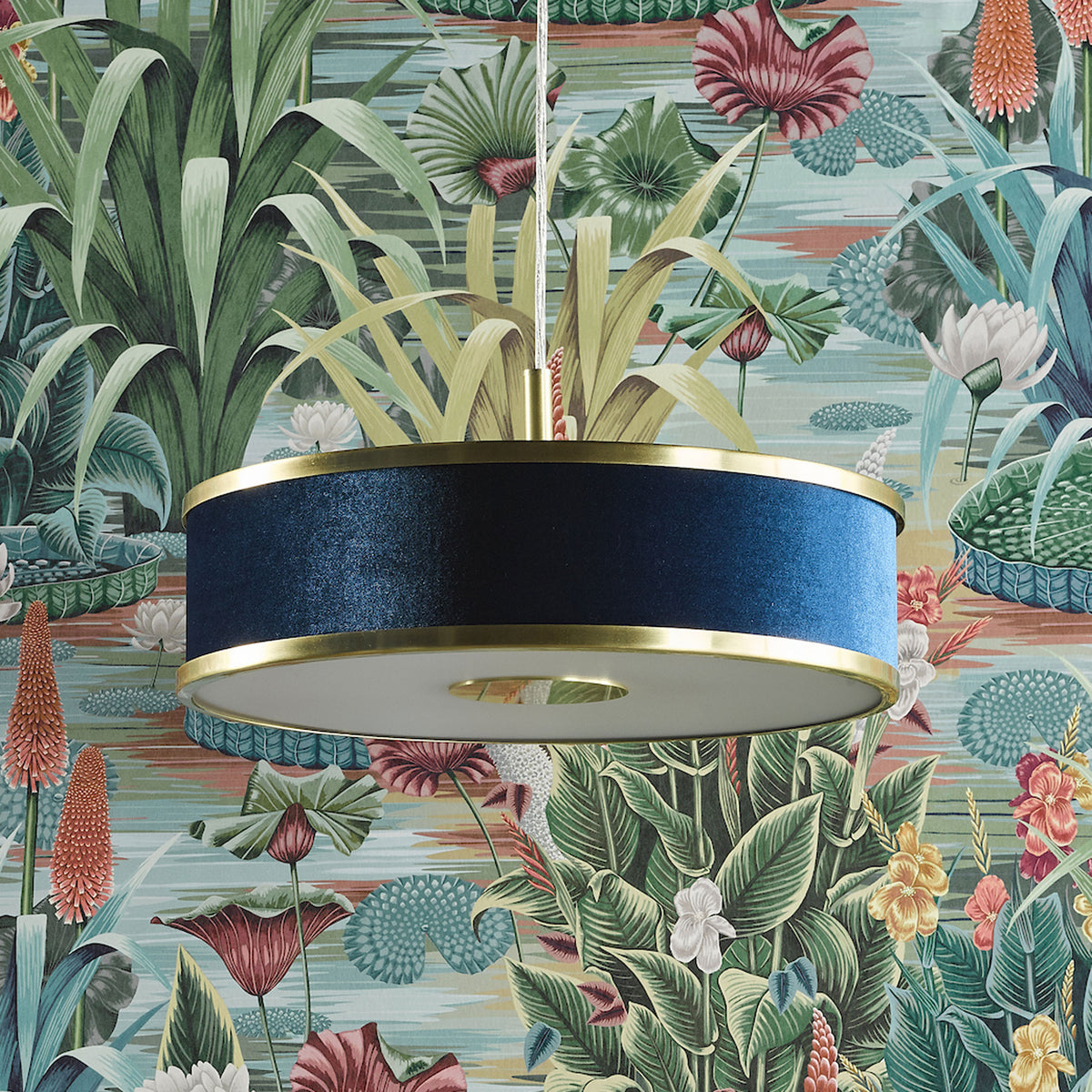 Blue velvet drum pendant light with gold trim against a tropical floral wallpaper.